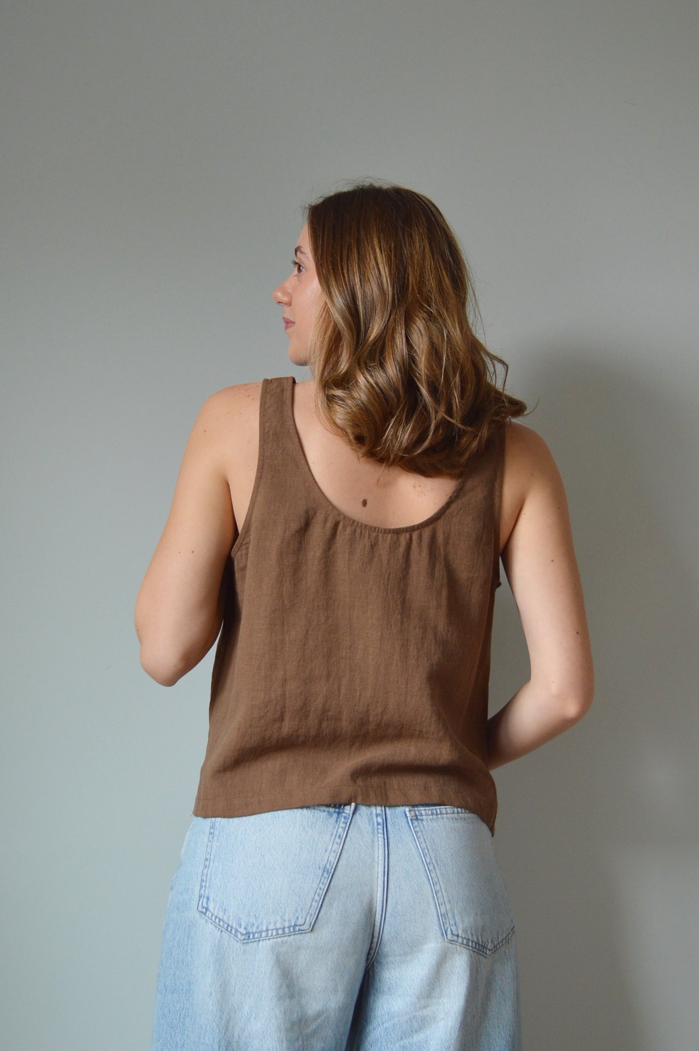 Woman in a brown tank top and blue jeans standing against a plain background