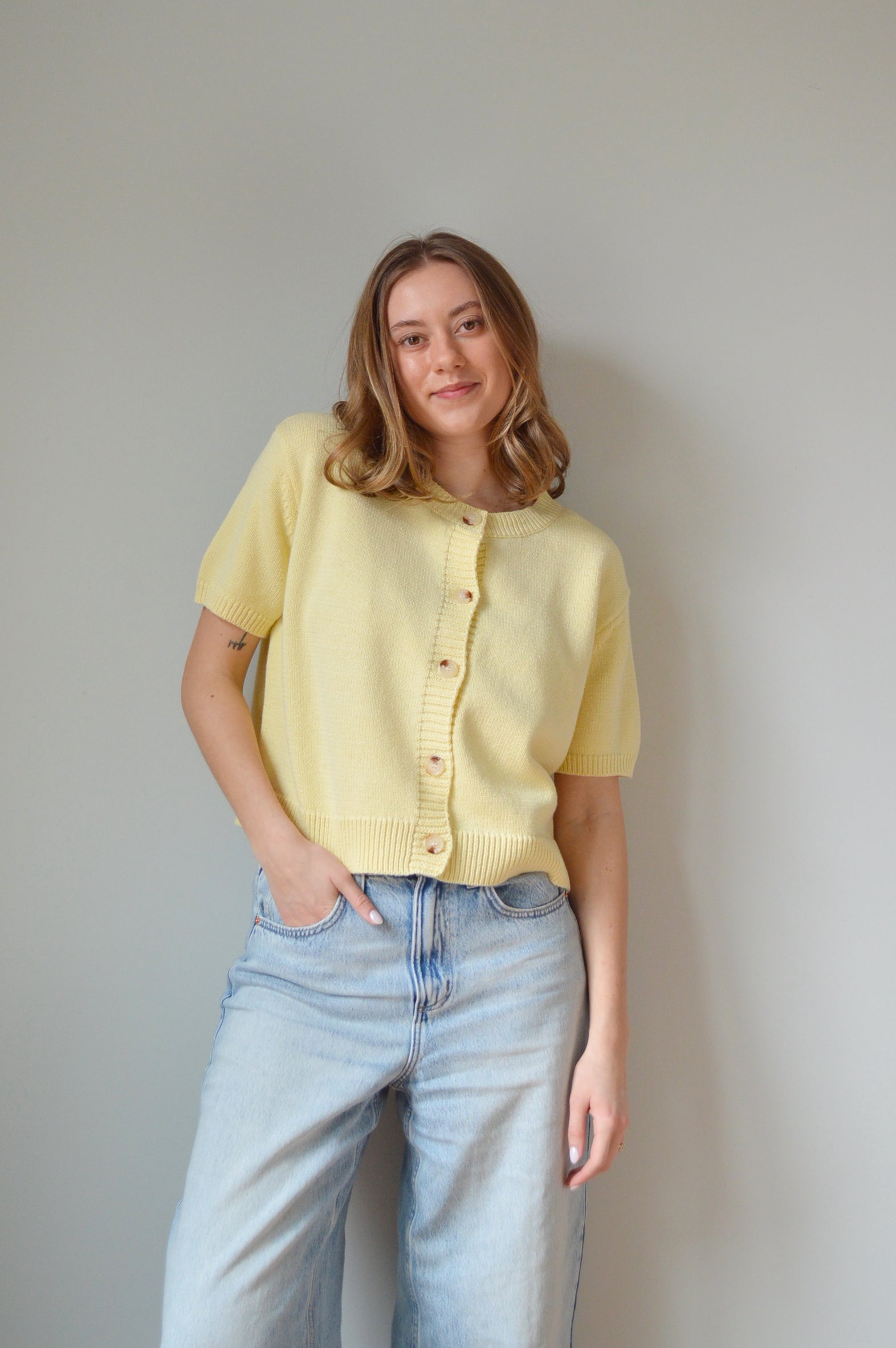 Woman wearing a yellow short sleeve cardigan with tan buttons and light blue jeans against a plain background