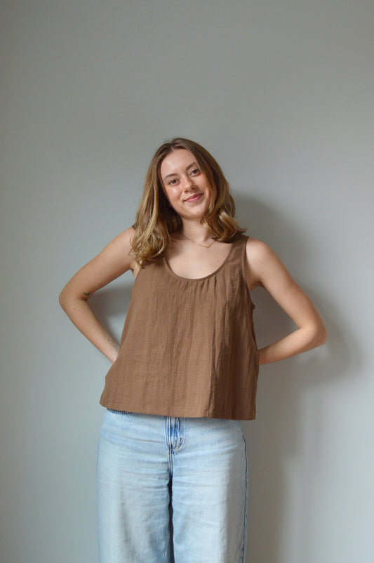 Woman in a brown tank top and blue jeans standing against a plain background