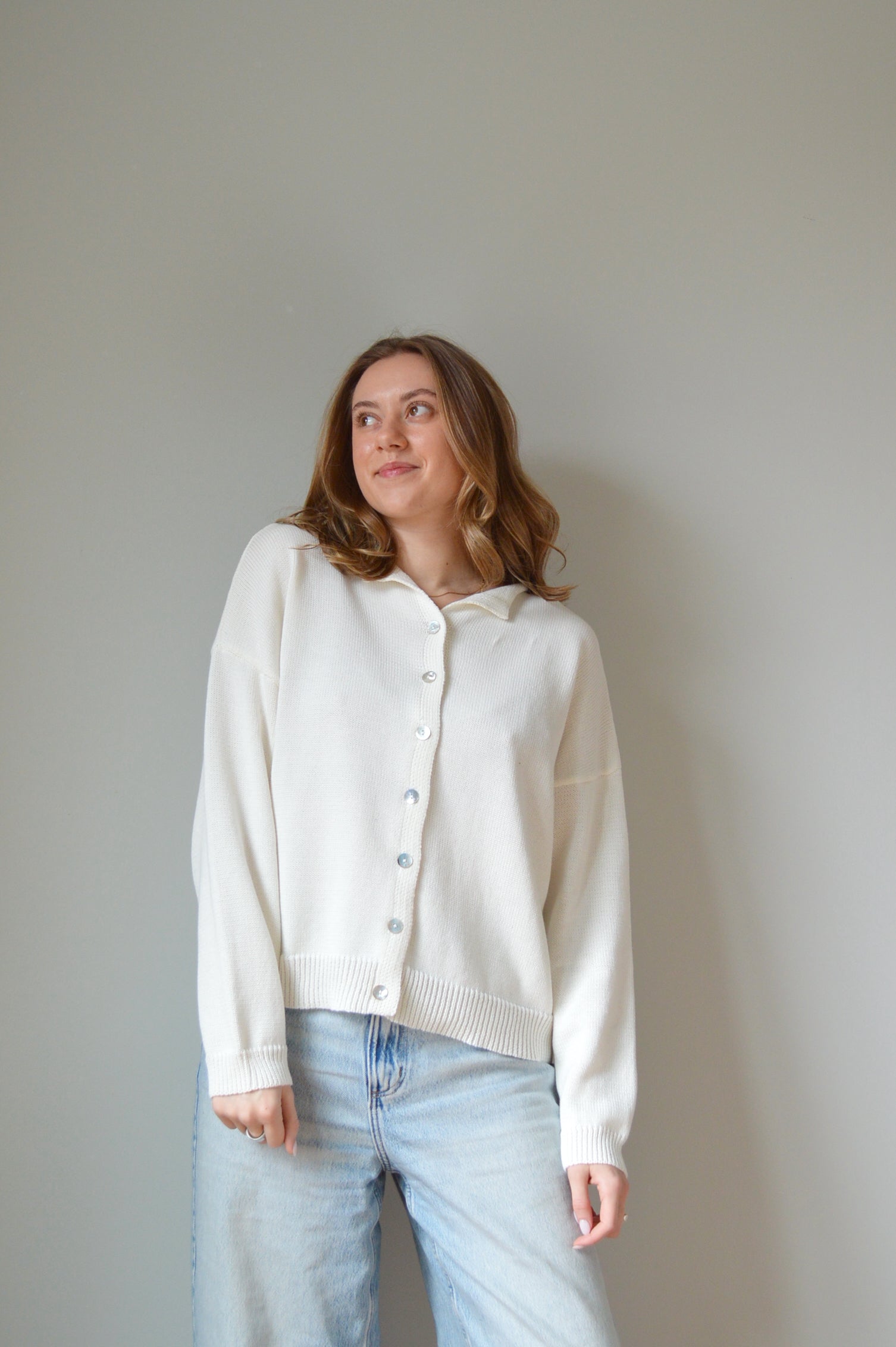 Person wearing a white sweater with pearl buttons and light blue jeans against a plain background