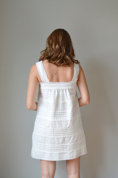 Woman wearing a white mini dress with pleated & eyelet detailing, square neck and buttons down the front against a plain background
