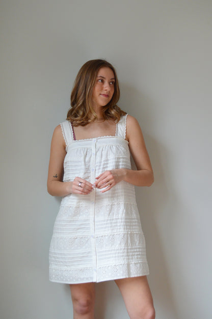 Woman wearing a white mini dress with pleated & eyelet detailing, square neck and buttons down the front against a plain background