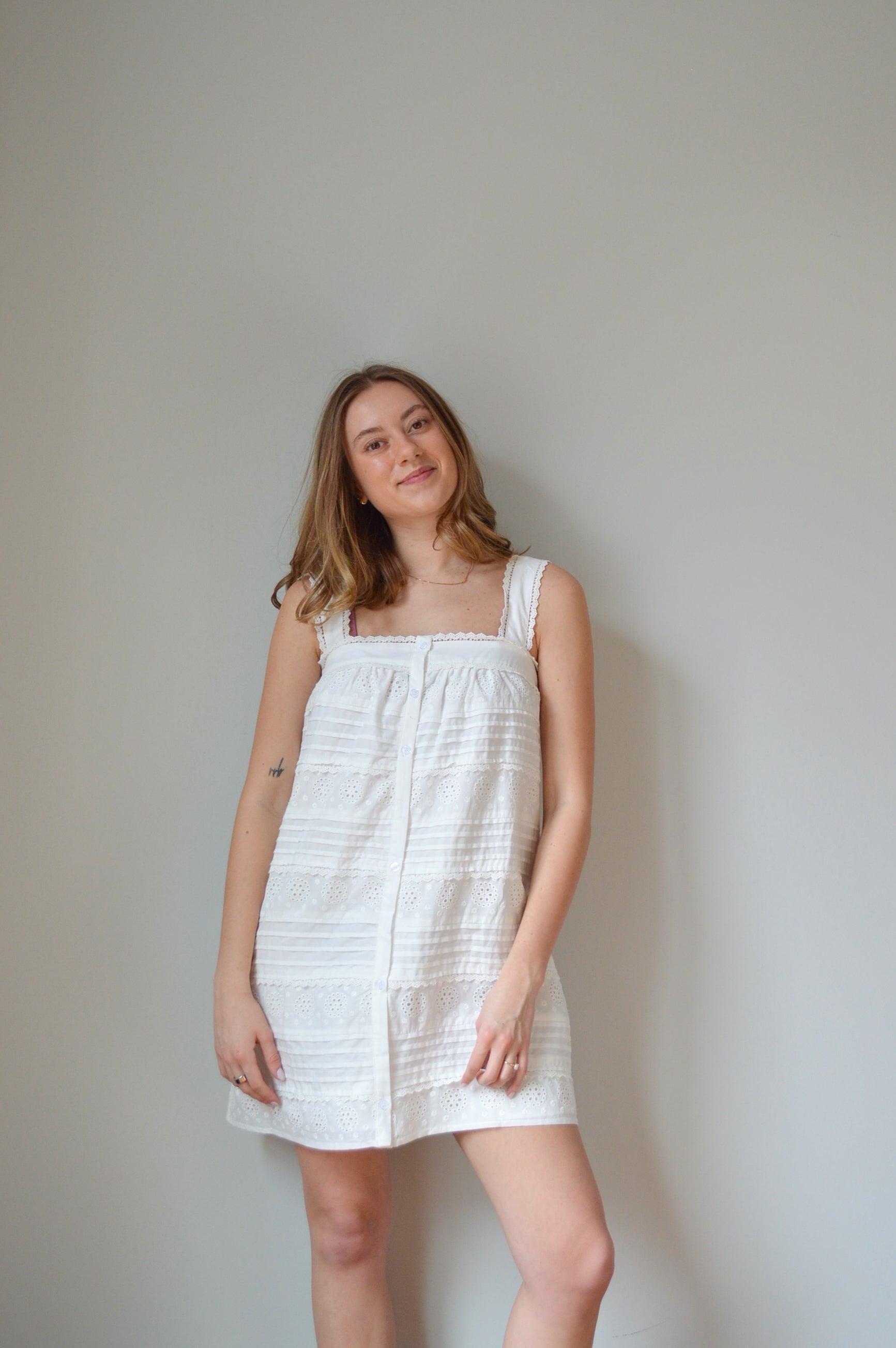 Woman wearing a white mini dress with pleated & eyelet detailing, square neck and buttons down the front against a plain background