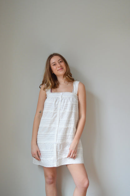 Woman wearing a white mini dress with pleated & eyelet detailing, square neck and buttons down the front against a plain background