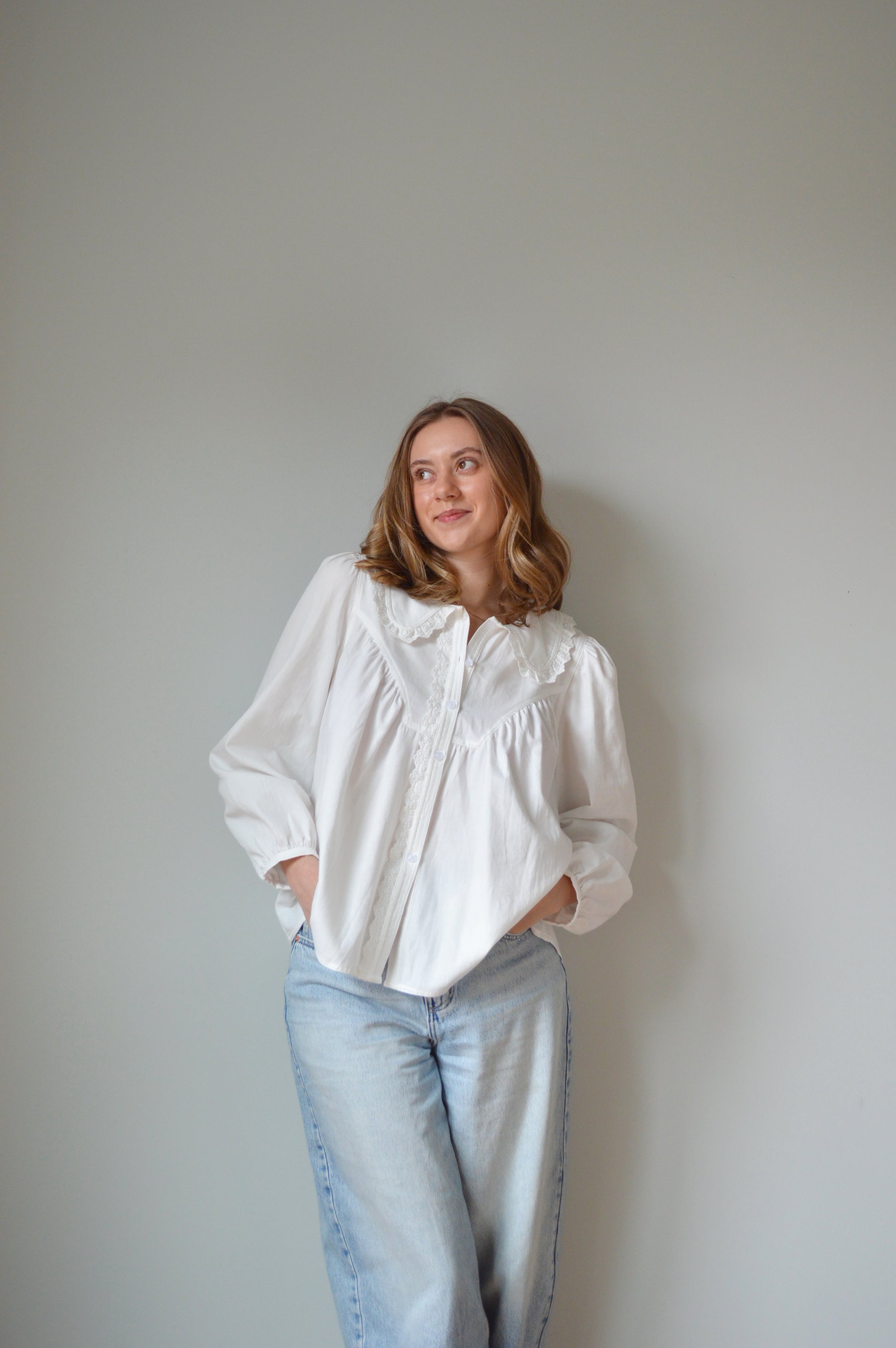 Person wearing a white button down shirt with a Peter Pan style collar, lace detailing and light blue jeans against a plain background