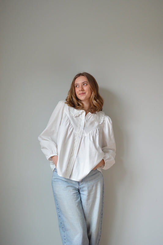 Person wearing a white button down shirt with a Peter Pan style collar, lace detailing and light blue jeans against a plain background