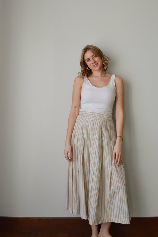 Woman wearing a white top and beige pleated skirt against a plain wall.