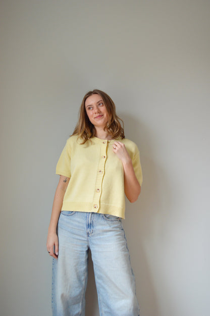 Woman wearing a yellow short sleeve cardigan with tan buttons and light blue jeans against a plain background