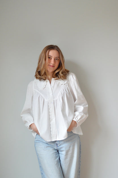 Person wearing a white button down shirt with a Peter Pan style collar, lace detailing and light blue jeans against a plain background