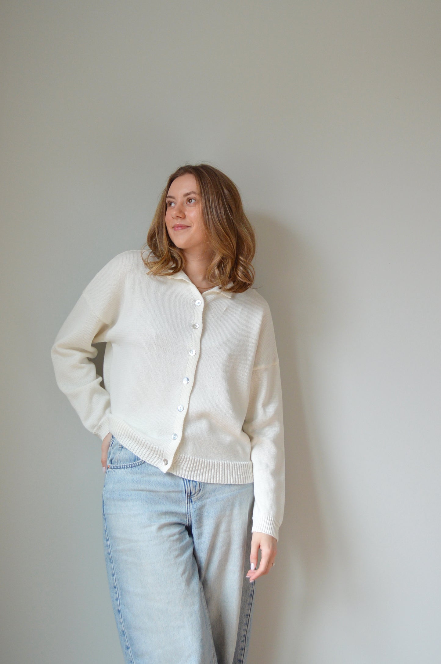 Person wearing a white sweater with pearl buttons and light blue jeans against a plain background
