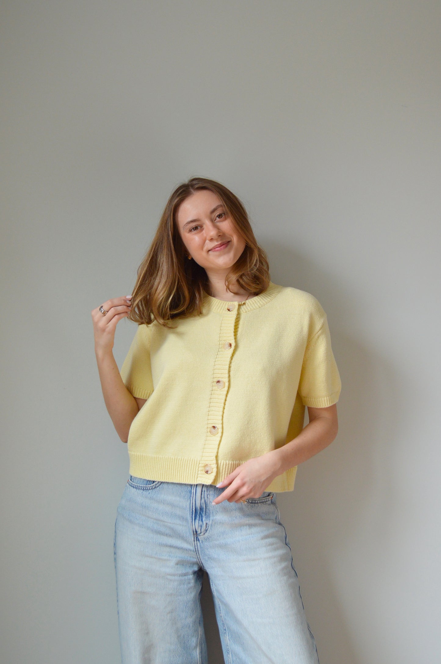 Woman wearing a yellow short sleeve cardigan with tan buttons and light blue jeans against a plain background