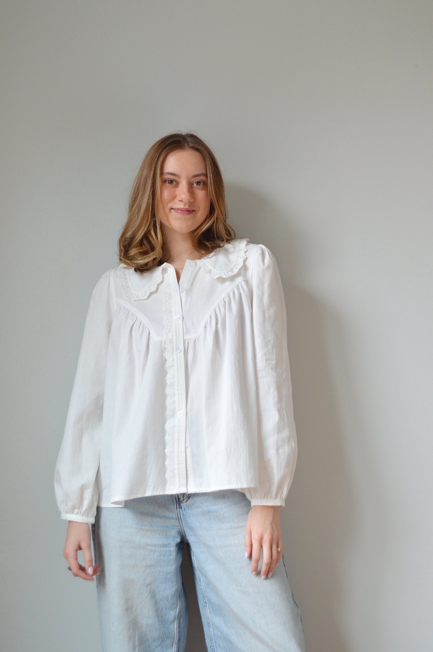 Person wearing a white button down shirt with a Peter Pan style collar, lace detailing and light blue jeans against a plain background