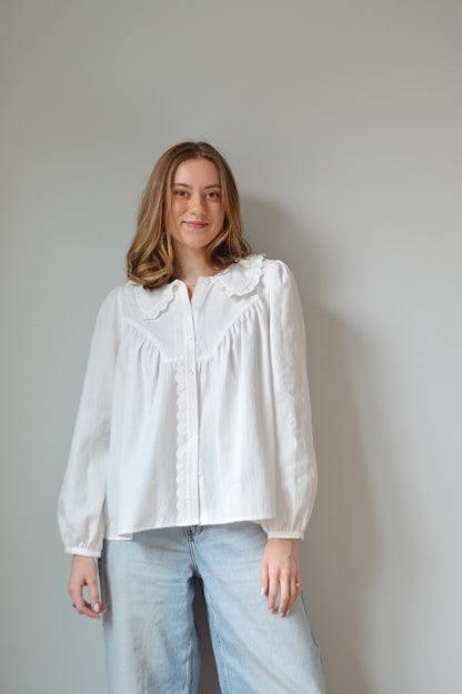 Person wearing a white button down shirt with a Peter Pan style collar, lace detailing and light blue jeans against a plain background