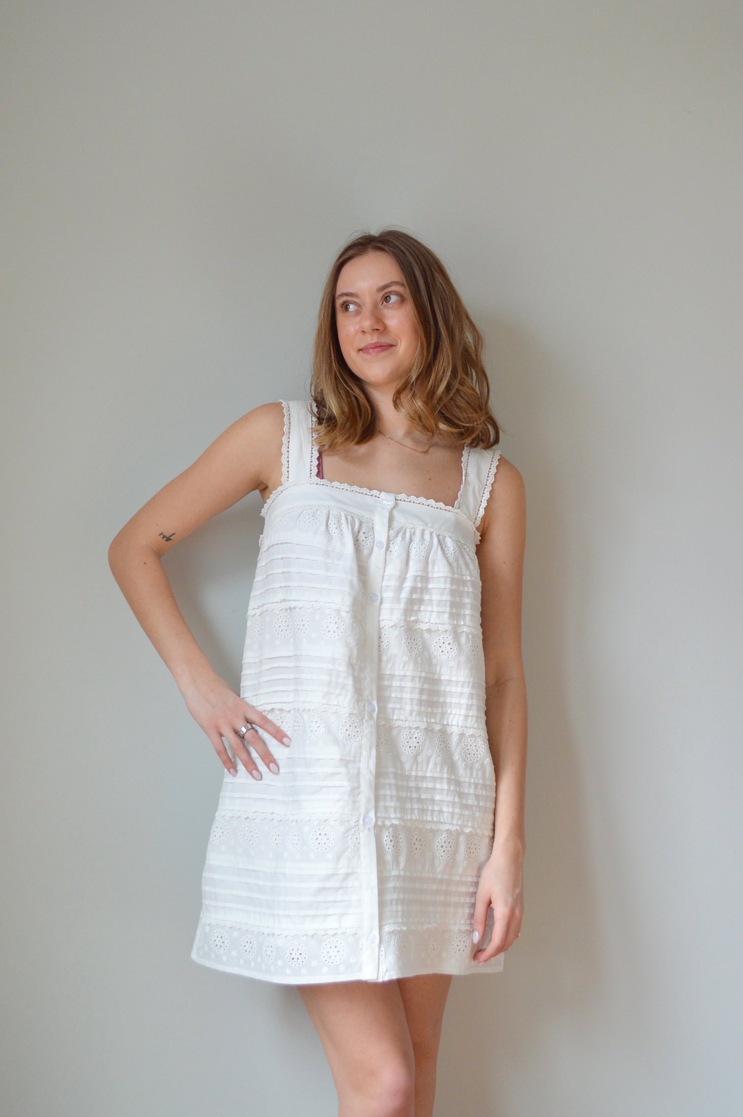 Woman wearing a white mini dress with pleated & eyelet detailing, square neck and buttons down the front against a plain background